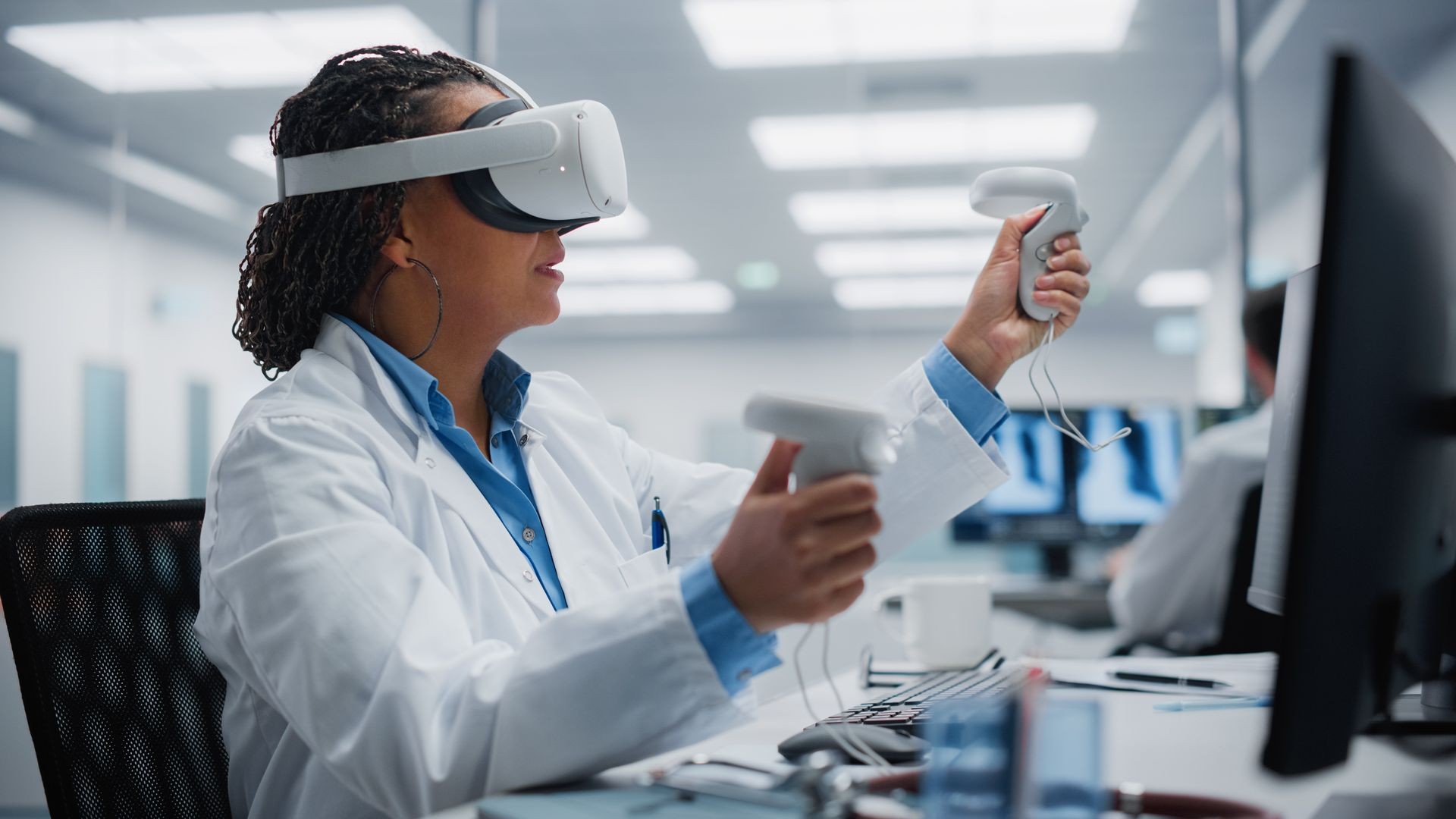 Person in a lab coat using a virtual reality headset and handheld controllers in an office setting.